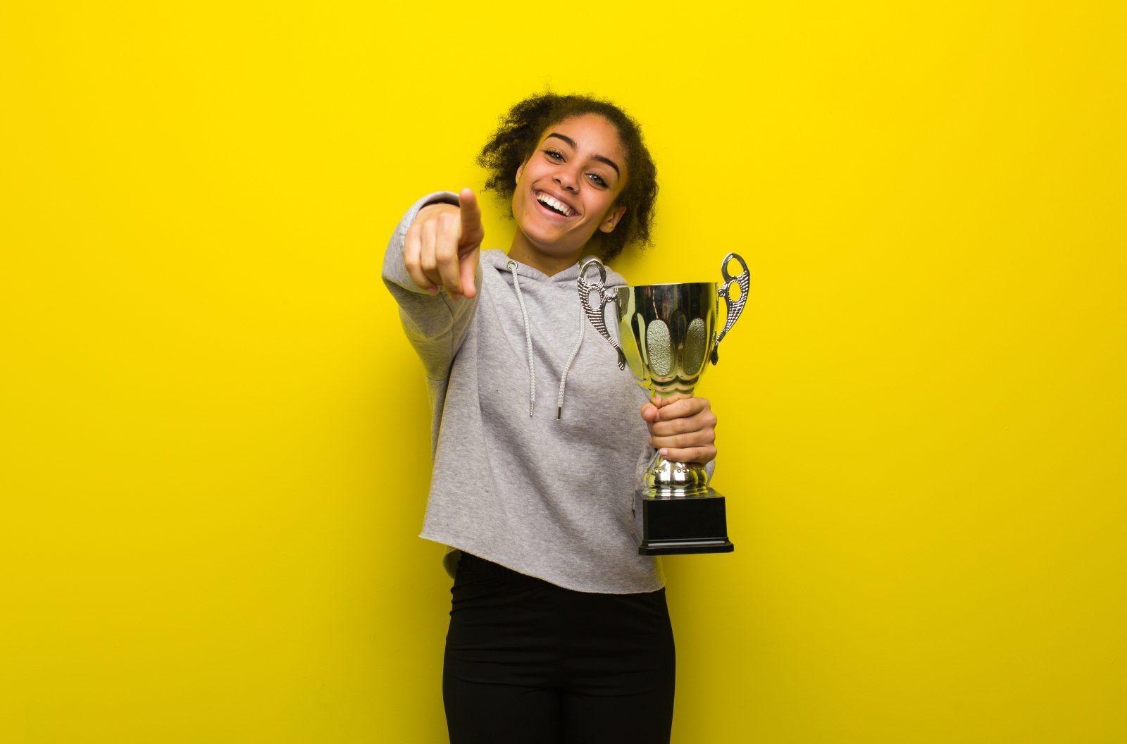 A young person holds a gold metal trophy while smiling and pointing toward the viewer.