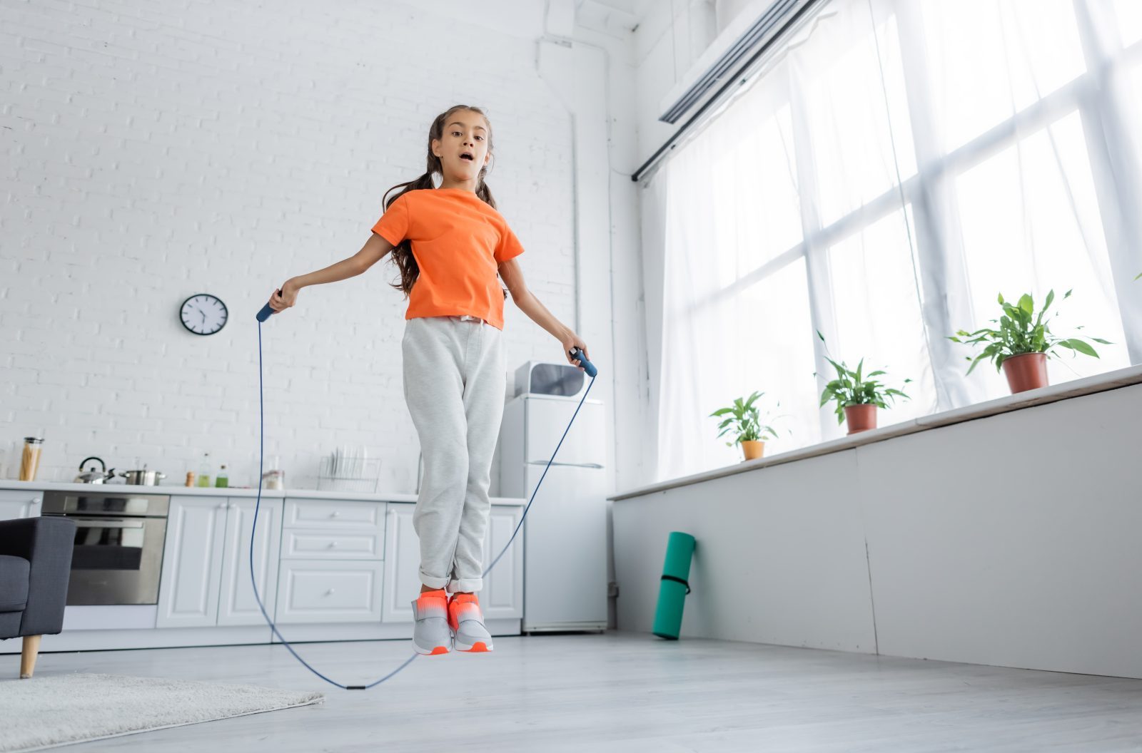 A girl in an orange shirt and sneakers jumps rope in a white living room.