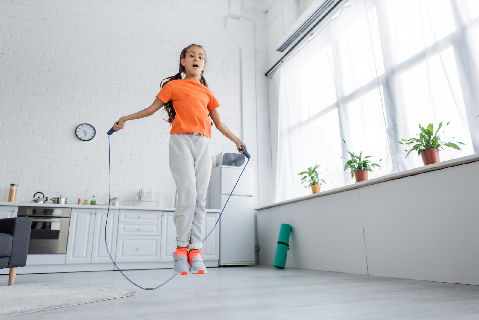 Una niña con una camisa naranja y tenis salta la cuerda en una sala blanca.