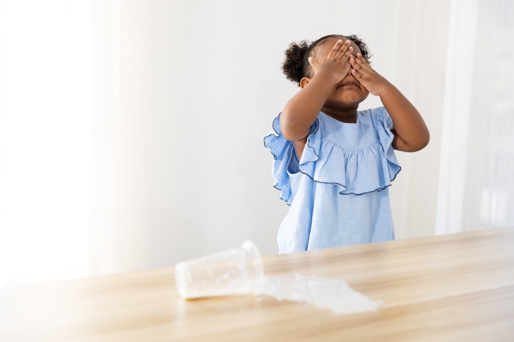 Milk spilled from a cup covers a kitchen table as a nearby child covers their face with their hands in frustration