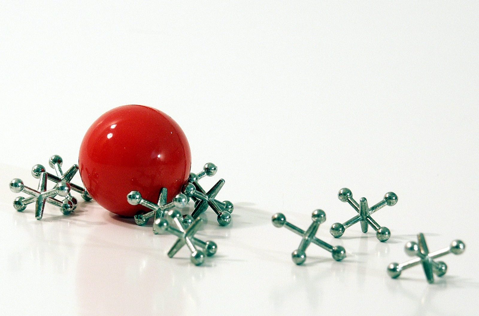 Una pequeña y brillante pelota roja junto a varios jacks de metal sobre un fondo blanco.