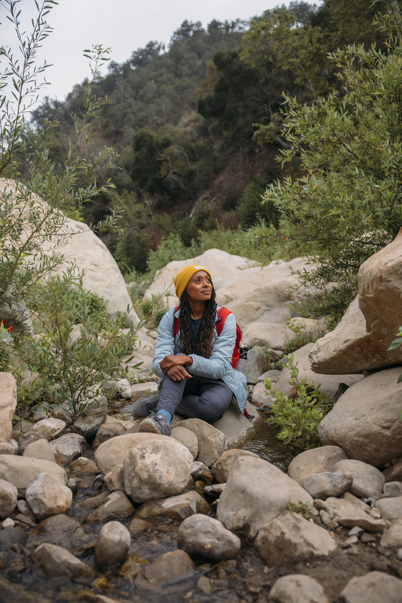A Black woman wearing a beanie sits amidst rocks in a natural outdoor setting. She looks up at the sky with a gentle smile.