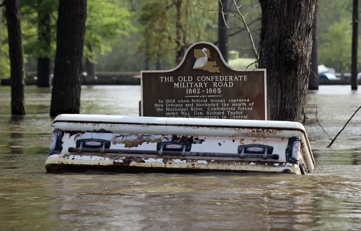 A white coffin lies sideways against a brown historical marker in deep water with trees surrounding it.
