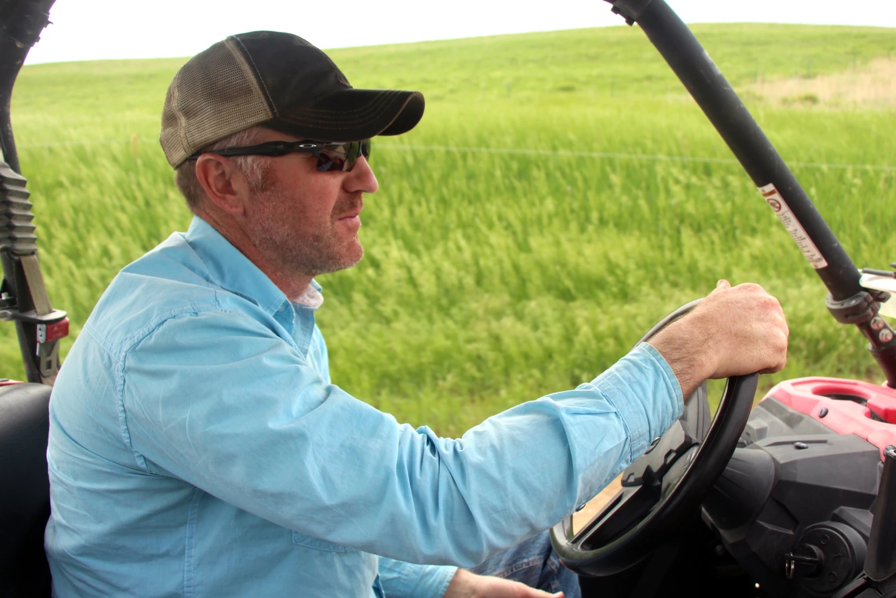 A man wearing a hat and sunglasses driving a vehicle on a prairie.