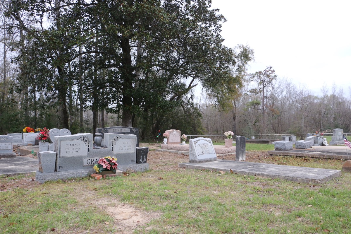 One headstone in a corner of a cemetery has grass in front of it, while others show above-ground vaults.  There is a large tree in the background.