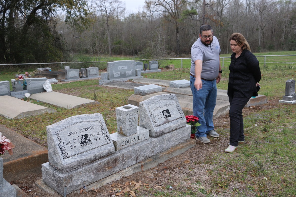 A man and a woman stand next to a double gravestone. He points to the grassy space in front of the grave as she looks on.