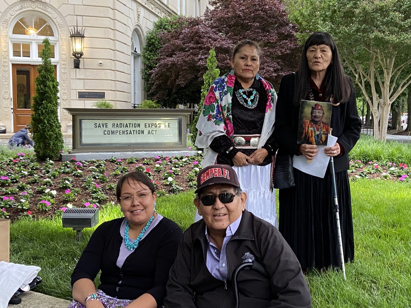 Four people, two sitting and two standing, in front of a sign that reads "Save Radiation Exposure Compensation Act"