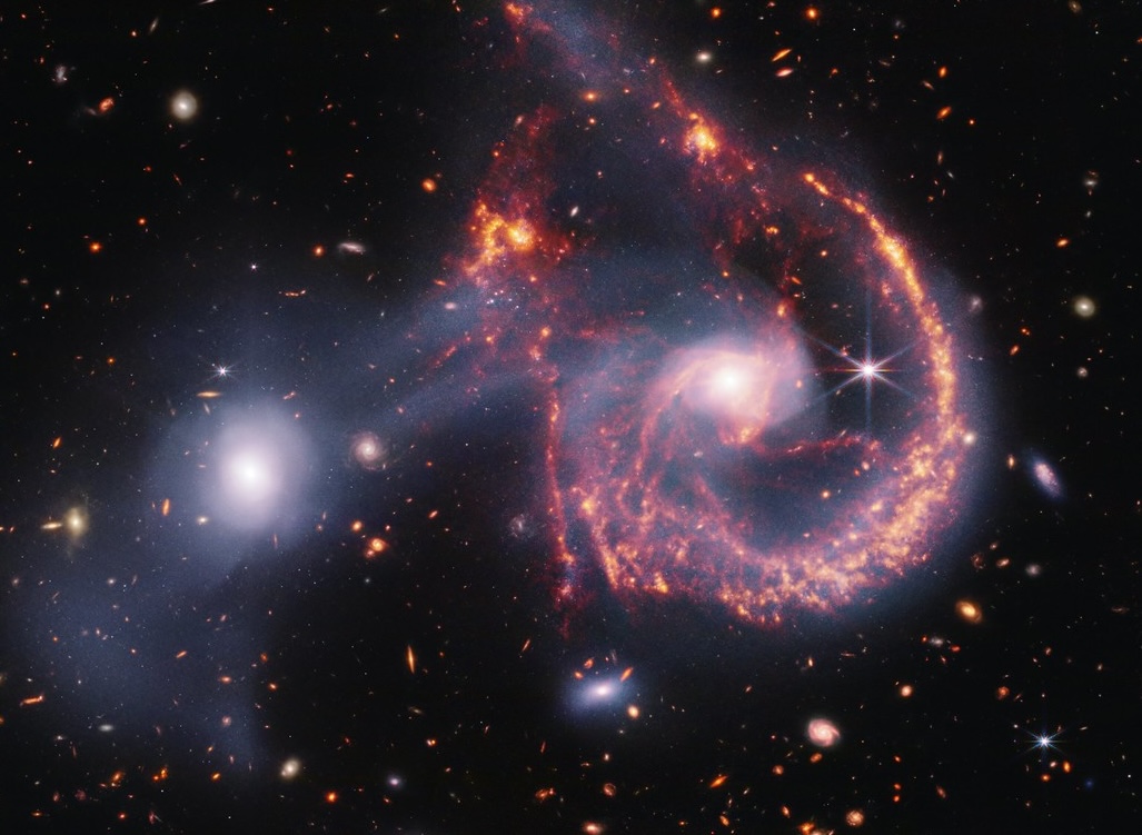 A pair of interacting galaxies. The larger of the two galaxies is slightly right of center, and composed of a hazy, bright, white center and a ring of gaseous filaments, which are different shades of red and orange. Toward the bottom left and bottom right of the ring are filaments of gas spiraling inward toward the core. At the top left of the ring is a noticeable gap, bordered by two large, orange pockets of dust and gas. The smaller galaxy to its left is made of hazy white gas and dust, which becomes more diffuse farther away from its center. To this galaxy’s bottom left, there is a smaller, more diffuse gas cloud that wafts outward toward the edges. Many red, orange, and white background galaxies are spread throughout, with some hazier in composition and others having more defined spiral patterns.