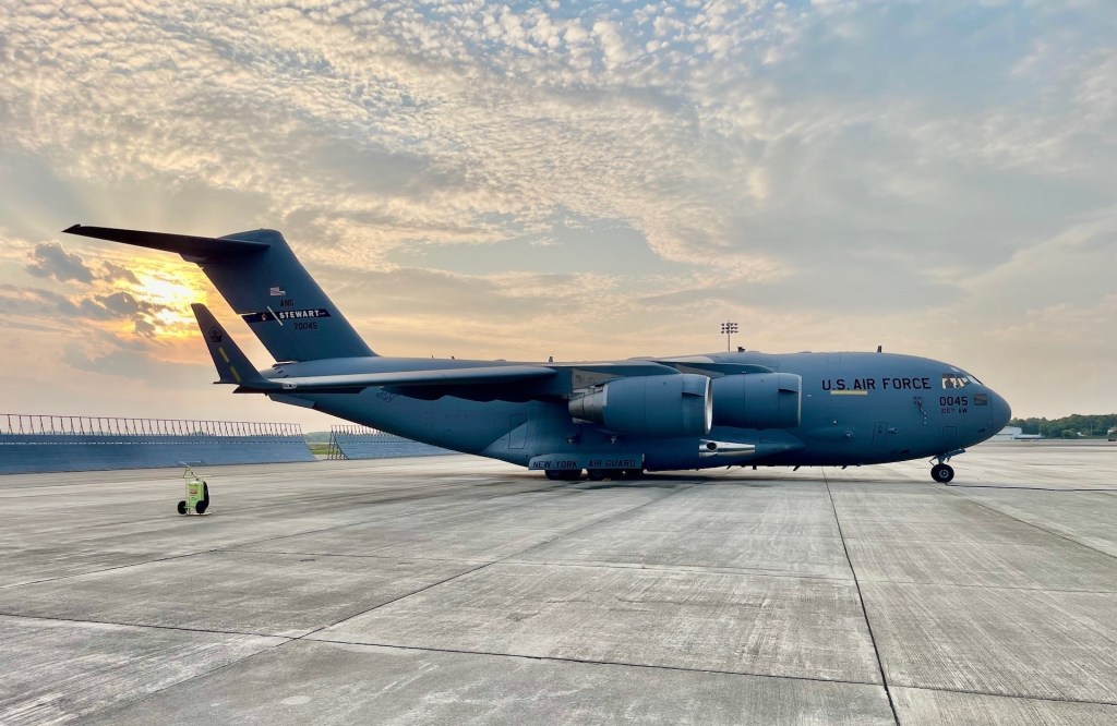 A huge, heavy gray aircraft sits on a white concrete tarmac with the sun setting in the background.