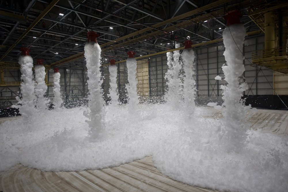Inside an empty hangar, white foam gushes down from several spouts long the ceiling, filling the floor with