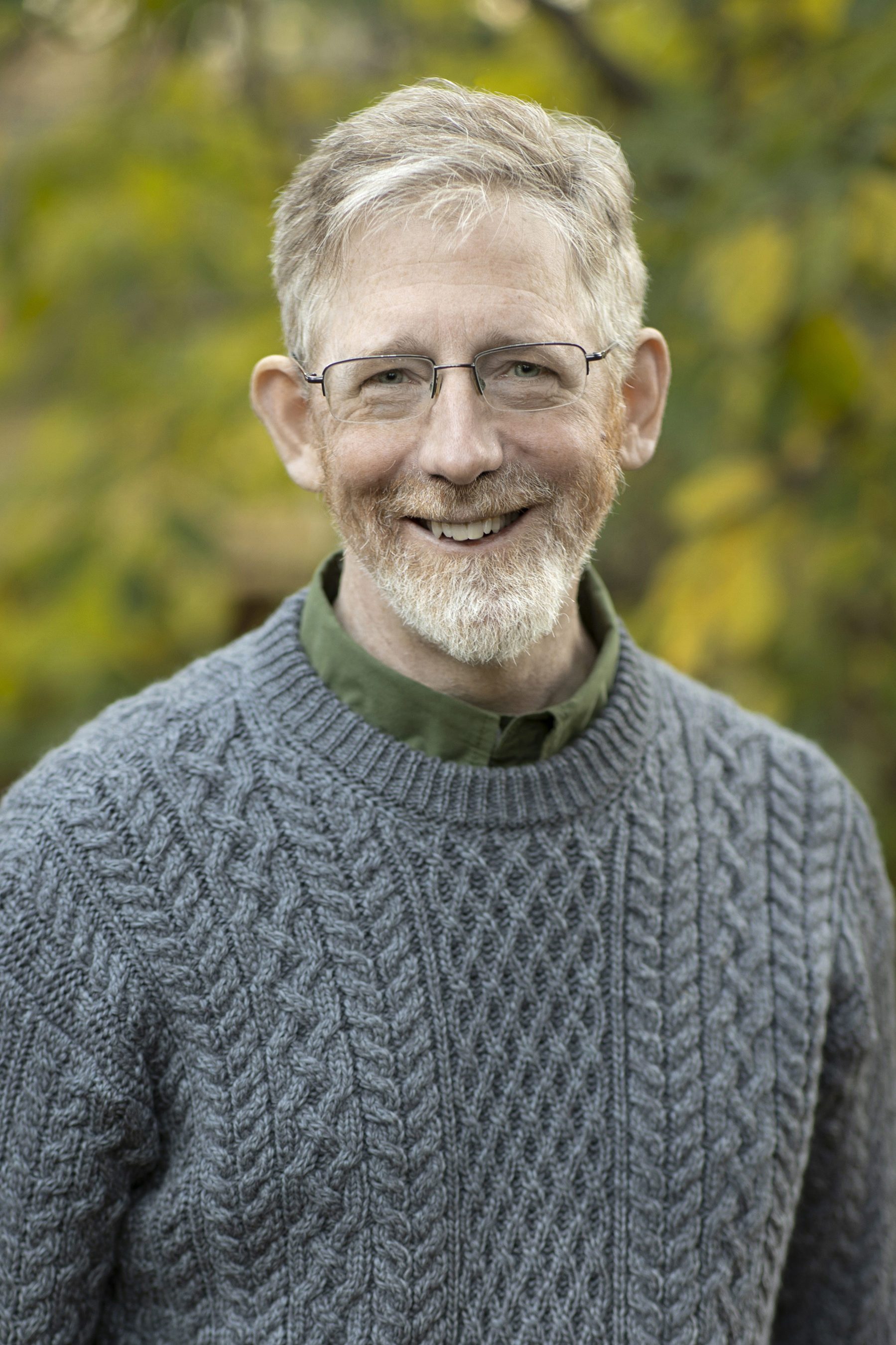 A smiling middle-aged man with gray hair and beard wearing glasses and a gray cable-knit sweater over a green collared shirt, photographed outdoors against a softly blurred background of autumn foliage in yellow and green tones.