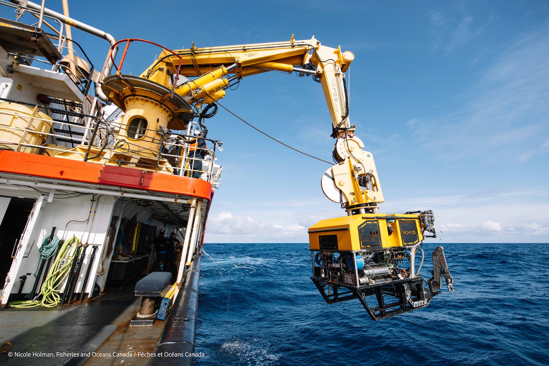 A blocky yellow and black machine hangs over the surface of the ocean from a crane arm attached to a ship.