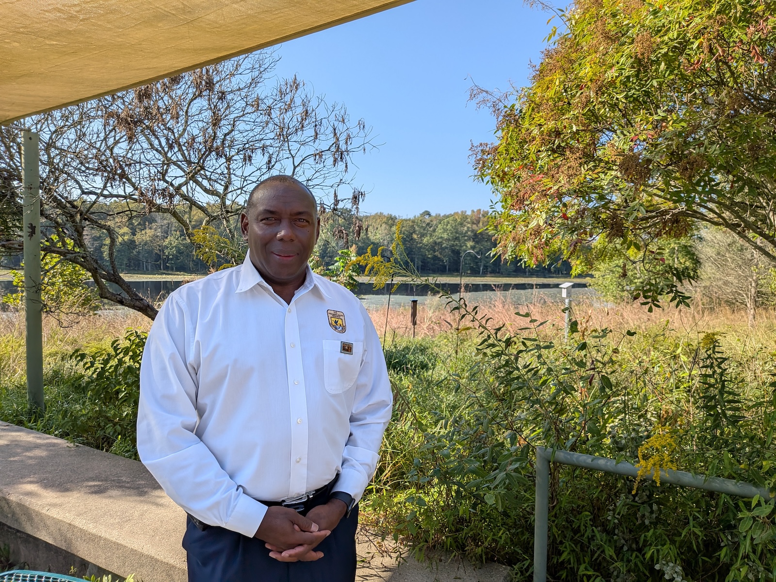 A man stands by a lake with lush vegetation overhanging it. There's a bird box behind him.