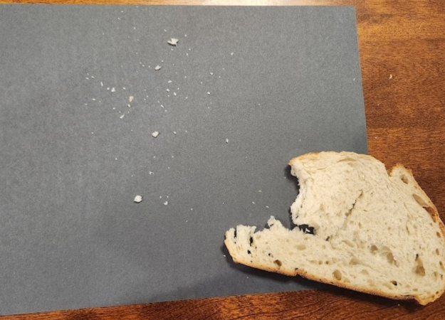 Bread crumbs scattered on black construction paper next to piece of artisanal bread on a wooden table.