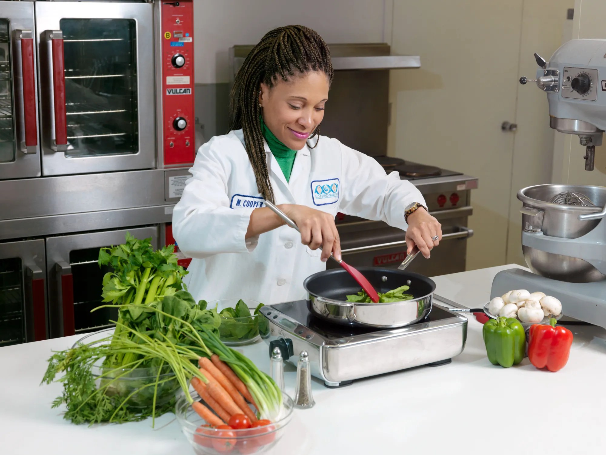 Chef in white coat and green shirt cooking in professional kitchen, stirring food in pan while surrounded by fresh vegetables including carrots, tomatoes, celery, green onions, leafy greens, bell peppers, and mushrooms.