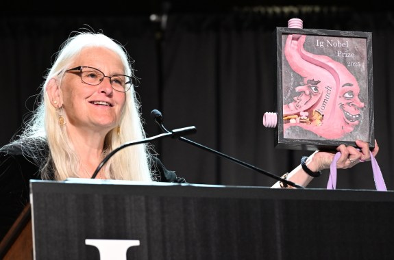 A woman holds up the 2025 Ig Nobel Prize- a portrait of the digestive system with funny faces on it.
