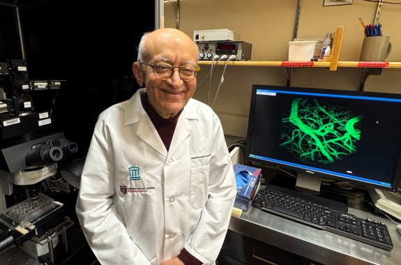 An older man smiling while standing next to a computer with a complex image of overlapping lines