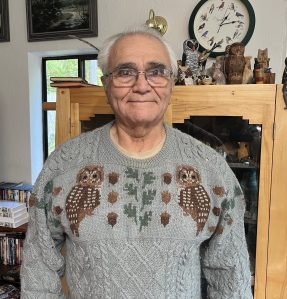 A senior man wearing glasses smiles while wearing a knitted sweater with a pattern of two spotted owls on it.
