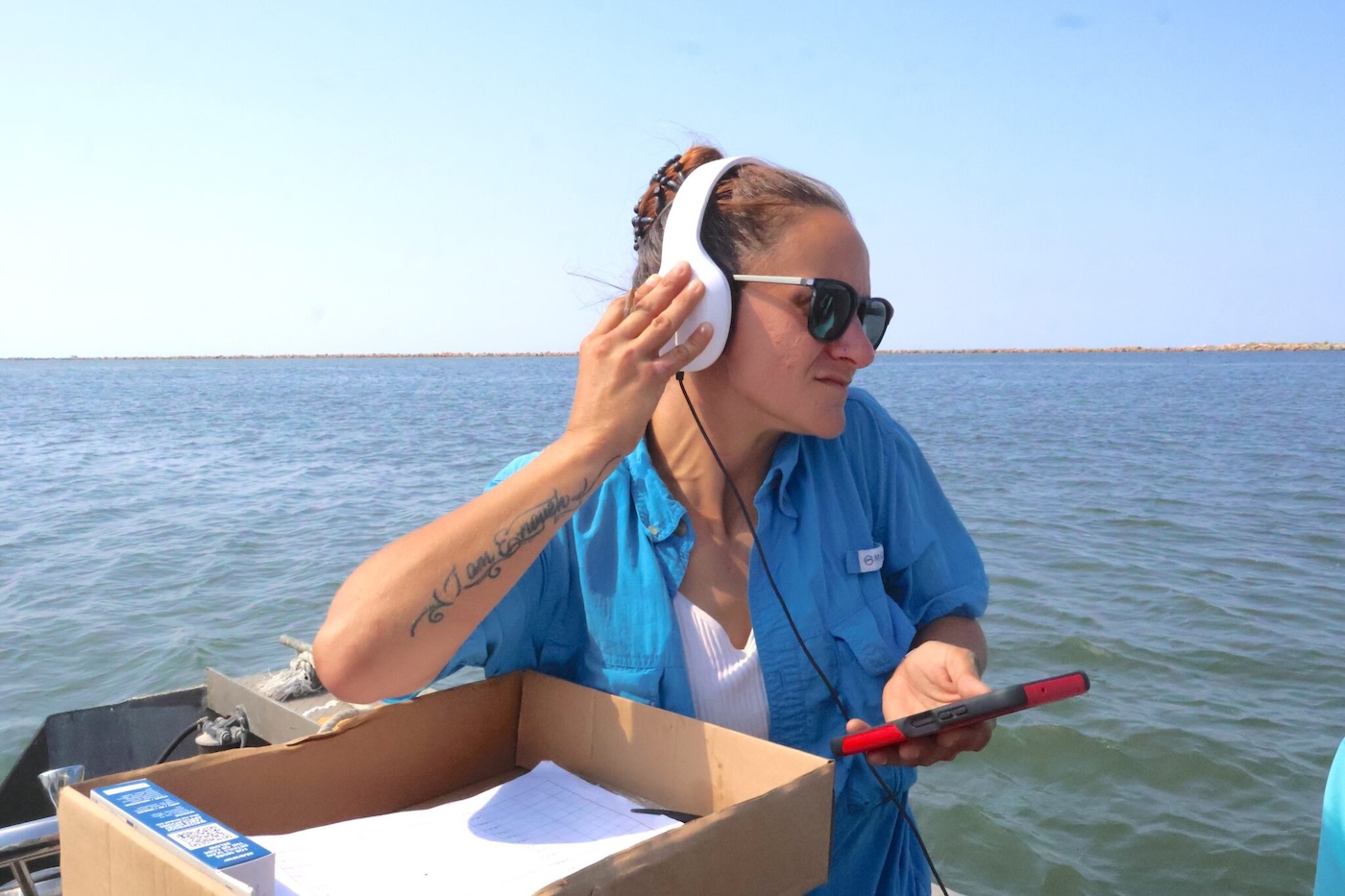 A woman in a blue shirt puts her hand up to her white headphones as she listens to something with the ocean in the background.