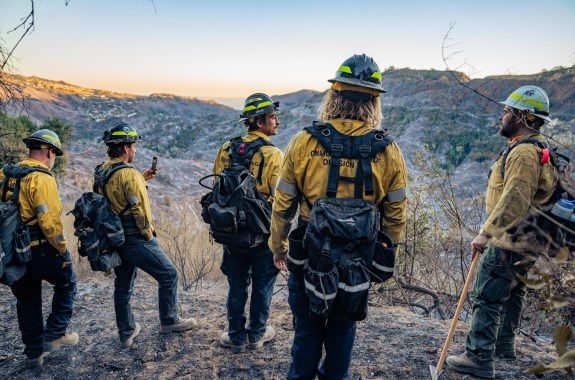 Five people in yellow jackets with large backpacks and helmets look out over a bare canyon.