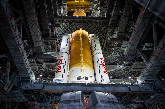 Looking up at a massive orange rocket standing with two white boosters, locked into a towering metal platform.