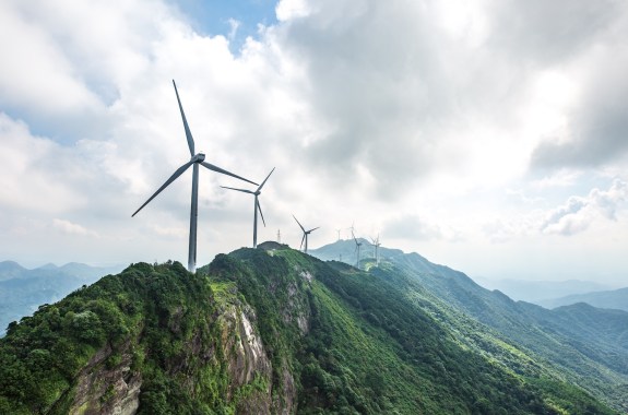 A row of windmills on a forested mountain ridge against a cloudy sky