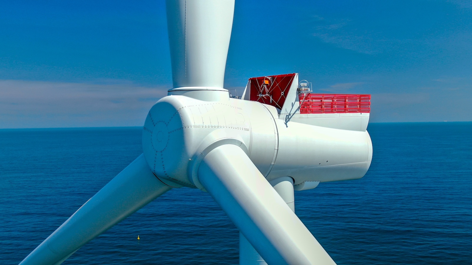 Close view of the center of a wind turbine, with three blades extending out of frame, and the ocean and horizon in the background
