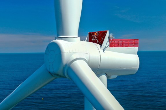 Close view of the center of a wind turbine, with three blades extending out of frame, and the ocean and horizon in the background