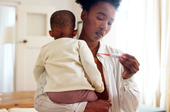 A woman holds a toddler in one arm and looks at a thermometer in her other hand, in a bright white room