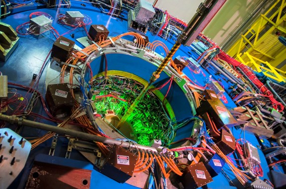Green light shines out of the center of a massive blue cylinder covered with colorful wires and boxes.