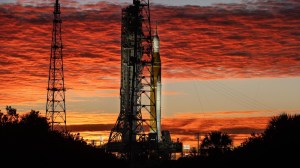 A rocket sits on a platform against bright orange clouds as the sun sets.