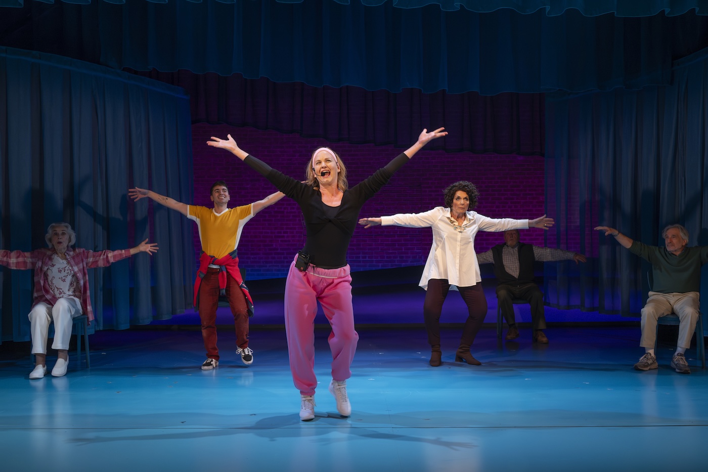 A woman in pink pants and a black shirt sings on stage with arms stretched up; performers in the background do the same.