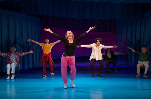 A woman in pink pants and a black shirt sings on stage with arms stretched up; performers in the background do the same.