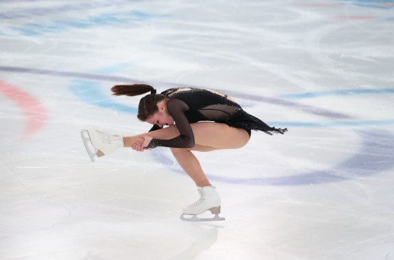A woman figure skater bends over her extended leg in a sit spin as her ponytail flies outward.