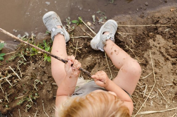 Looking straight down on a blond toddler sitting in the dirt at the edge of water with legs extended.