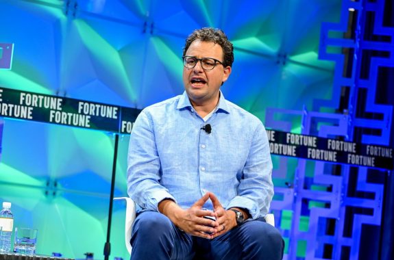 A man in a blue shirt sits in front of a blue and green background with the word "Fortune" running across it. He wears glasses, and he's speaking with his hands held together in his lap.