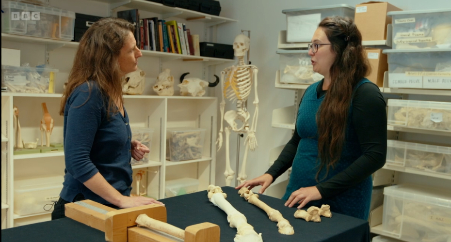 Two women talk while standing in a room in which clear bins of bones are stored on shelves. Several skulls and a full-sized human skeleton are in the background. Several horse leg bones are displayed on a table in front of the women.