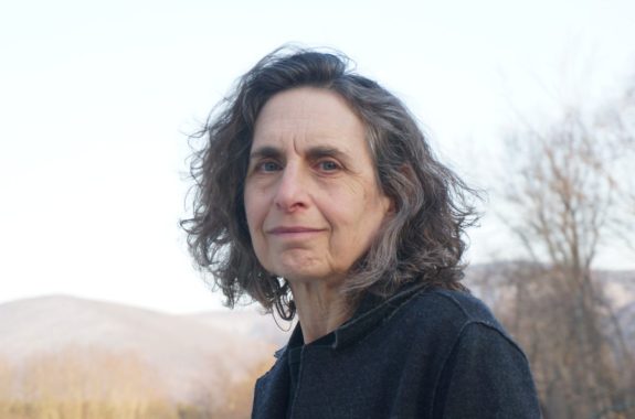 A woman with gray, shoulder-length hair looks at the camera. A faded background of bare trees and hills is behind her.