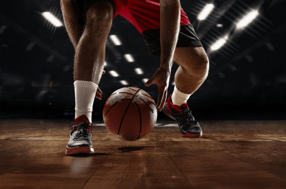 A close up of a basketball player dibbling a ball between his legs, on a wooden arena floor with a dark background