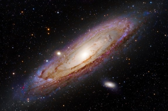 An oblong, cloudy whitish and purplish galaxy, slanted against a starry black sky.