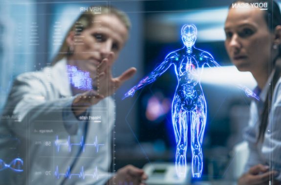 Young female physician and young female patient analyze transparent screen in front of them, depicting an outline of an AI-generated human form in blue.