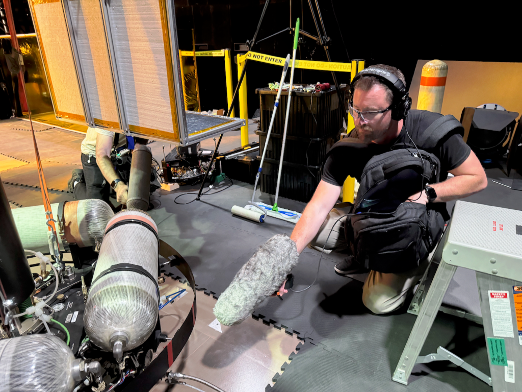 A man wearing headphones squats near a mechanical device with tanks, holding out a long furry microphone.