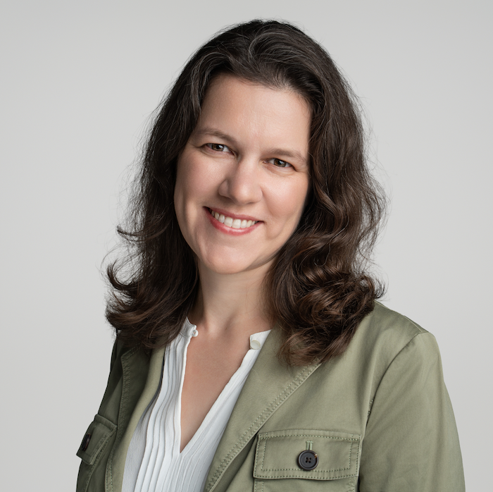 A woman with wavy brown hair and an olive green jacket smiling.