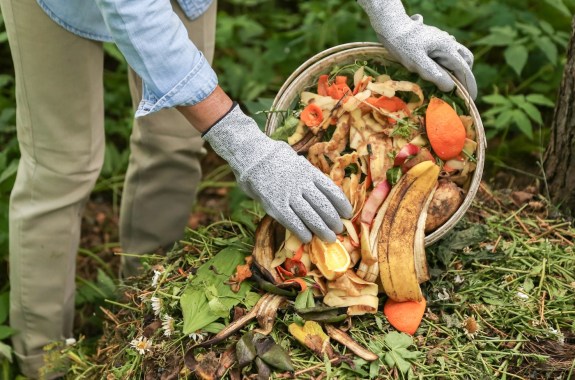 Close-up of gloved hands dumping a bucket of kitchen scraps into a pile of green and brown vegetation.