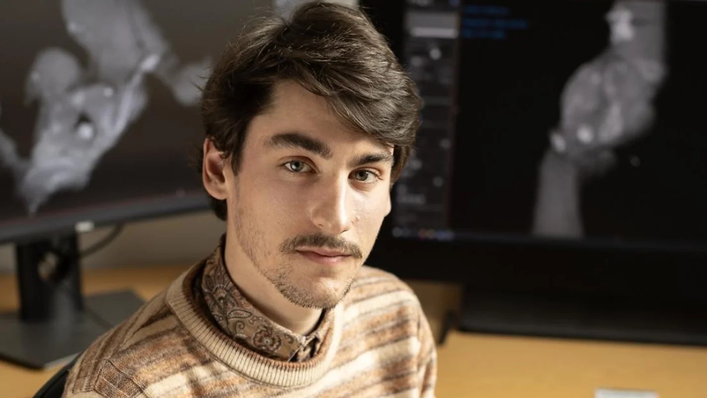 A young man with dark hair and a mustache wearing a striped brown sweater. Behind him sit two computer screens with images of fossils.