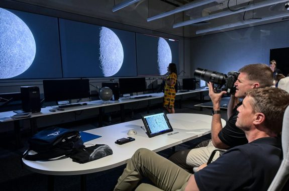 Two men sit behind a desk while one points a camera at images of the moon on large monitors across the room