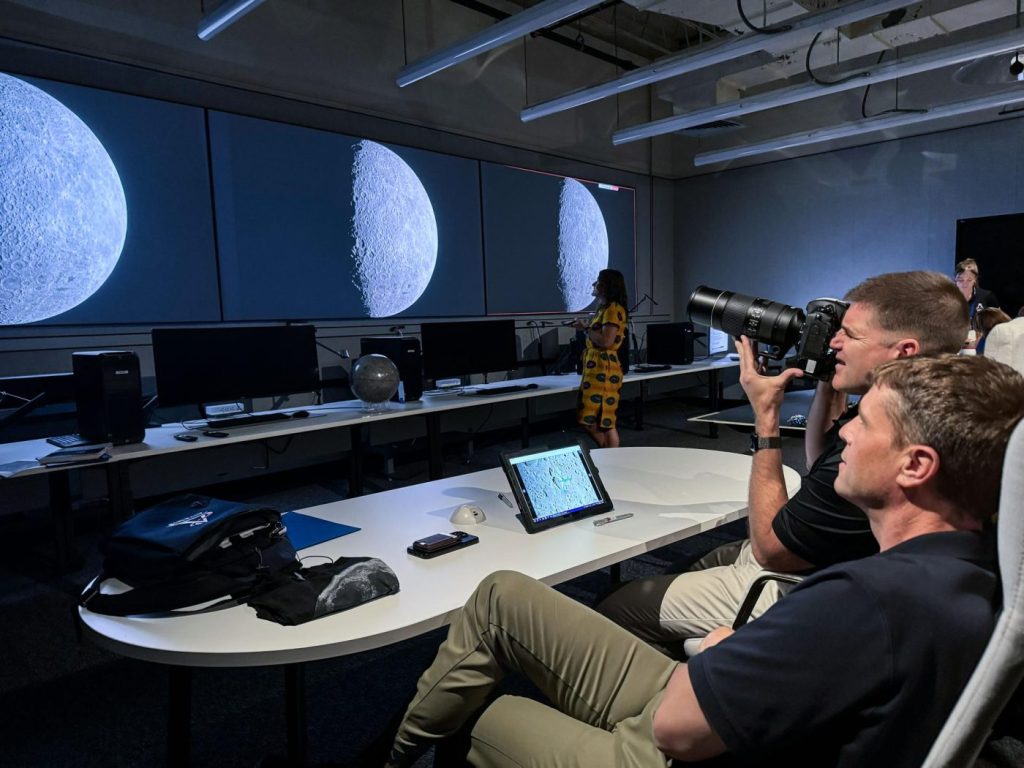 Two men sit behind a desk while one points a camera at images of the moon on large monitors across the room