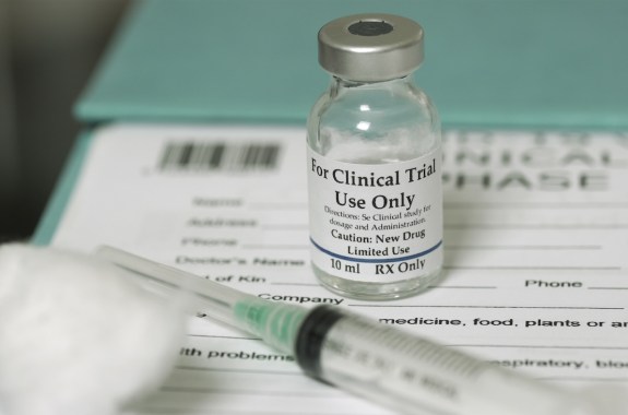 A small vial labeled "for clinical trial use only" sits next to a syringe and a cotton ball on top of a patient form.