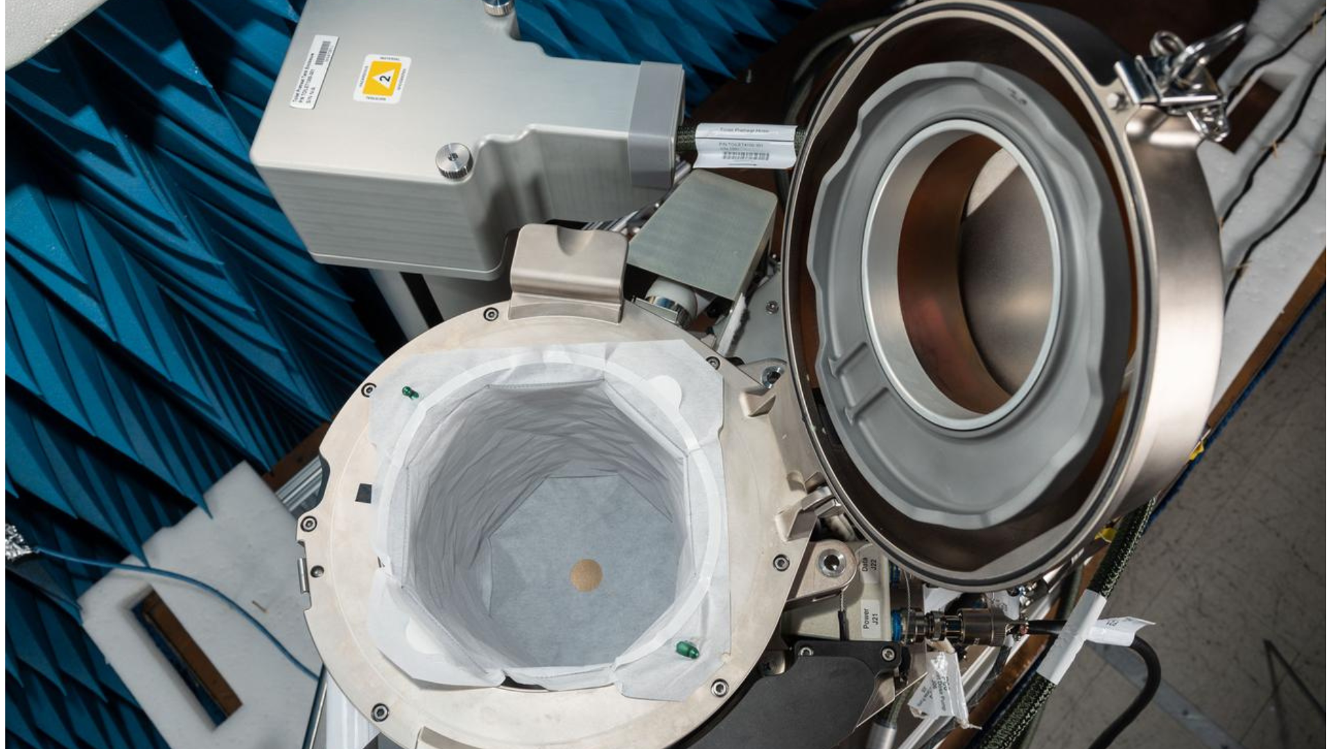 A top-down view of the ISS Universal Waste Management System (UWMS), a compact space toilet, shown with its lid open during preflight testing. A white collection bag lines a metal circular opening.