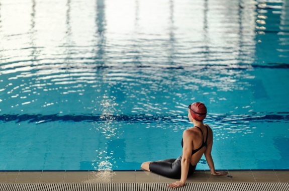 Rear view of a swimmer girl sitting on pool edge and looking at water.